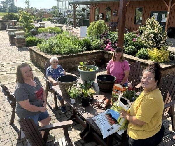 Four women smiling and gardening together at an outdoor table on a sunny day.