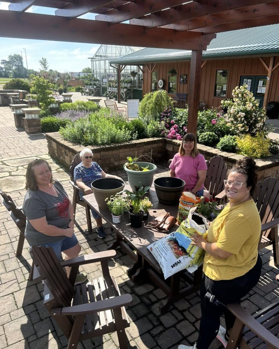 Four women smiling and gardening together at an outdoor table on a sunny day.