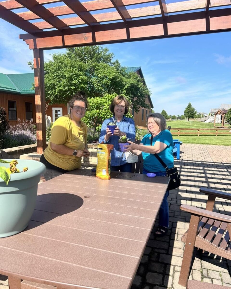 Three women potting plants together at an outdoor table under a wooden pergola.